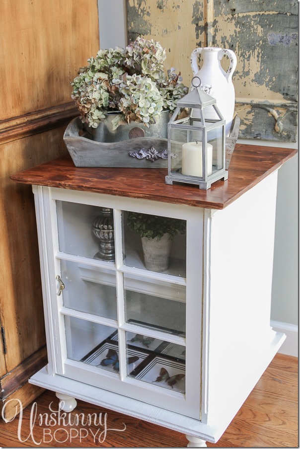 This end table is decorated with rustic pieces that reflect a farmhouse style, like the flowers in these vintage steel watering cans, the white ceramic vase, and a gilded metal lantern. 