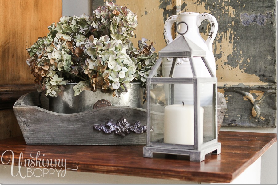 A closer look at the vintage and rustic table decorations. The metal candle lantern and metal watering cans holding flowers give this end table a uniquely farmhouse style. 