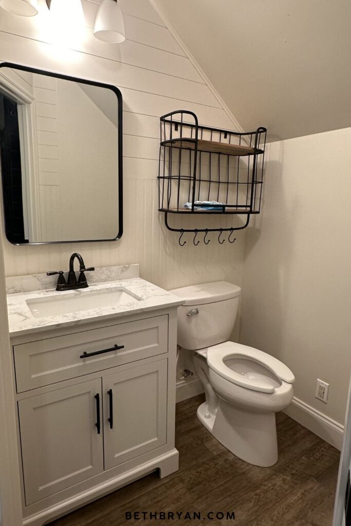 finished attic half bathroom with white shaker vanity matte black faucet and hardware shiplap wainscoting vanity light above black framed mirror and wire basket wall shelf under sloped roofline